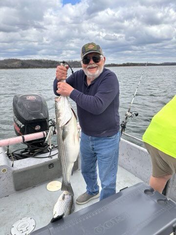 A smiling person holds a large fish while standing on a boat on a lake under a cloudy sky.