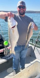 A smiling person in a hat and sunglasses stands on a boat on the water, holding up two large striped fish.
