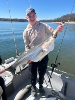 A person holding a large striped bass on a boat on a sunny day with calm water in the background.