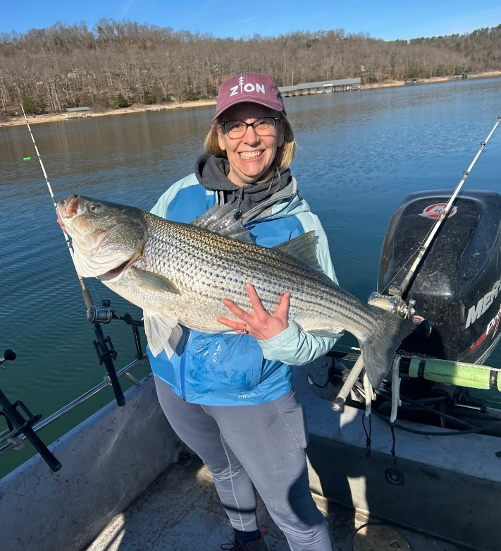Woman on boat holds large striped bass, blue water and trees in the background. She's smiling, wearing glasses and a hat.