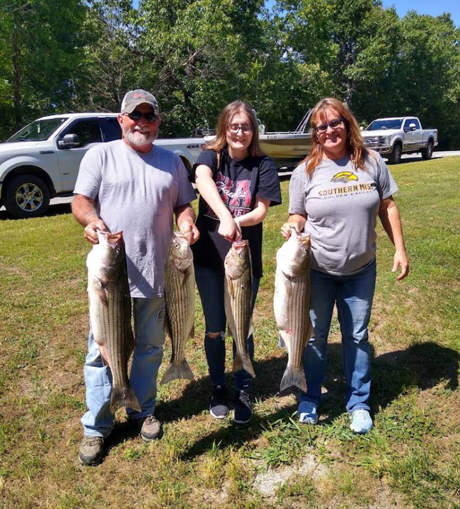 Three people holding up large striped bass they caught, outdoors.