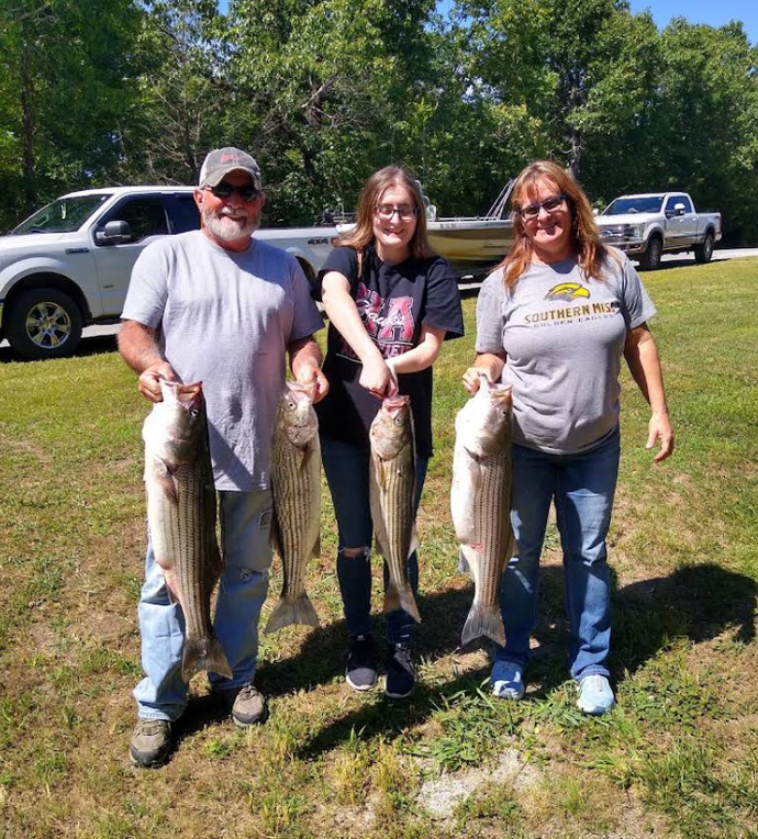 Three people holding up large striped bass they caught, outdoors.