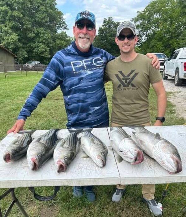 Two men pose with a table of caught fish outdoors. They smile; one wears a fishing shirt, the other a green shirt.