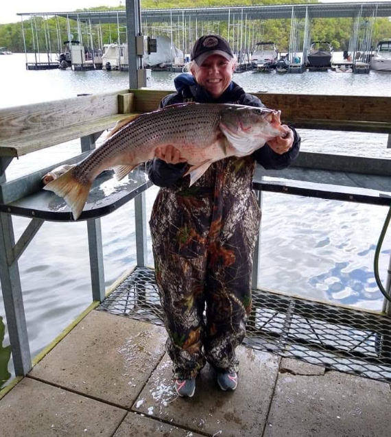 Woman in camo waders holds large striped bass at a dock.