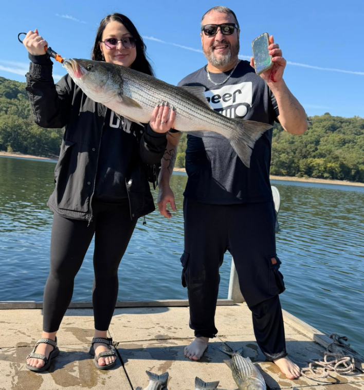 Two people holding a large striped fish on a dock, near water and green hills.