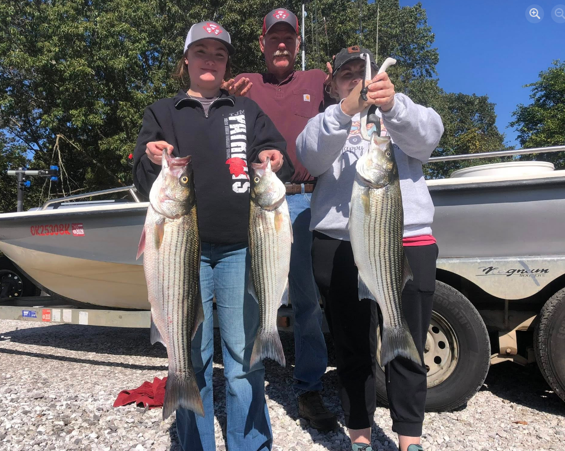 Three people hold up striped bass caught on a boat, standing on gravel in front of water and a blue sky.