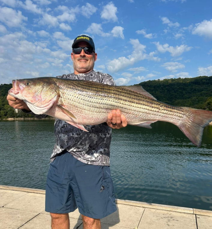 Man holding a large striped bass he caught near a body of water, wearing sunglasses and a cap.