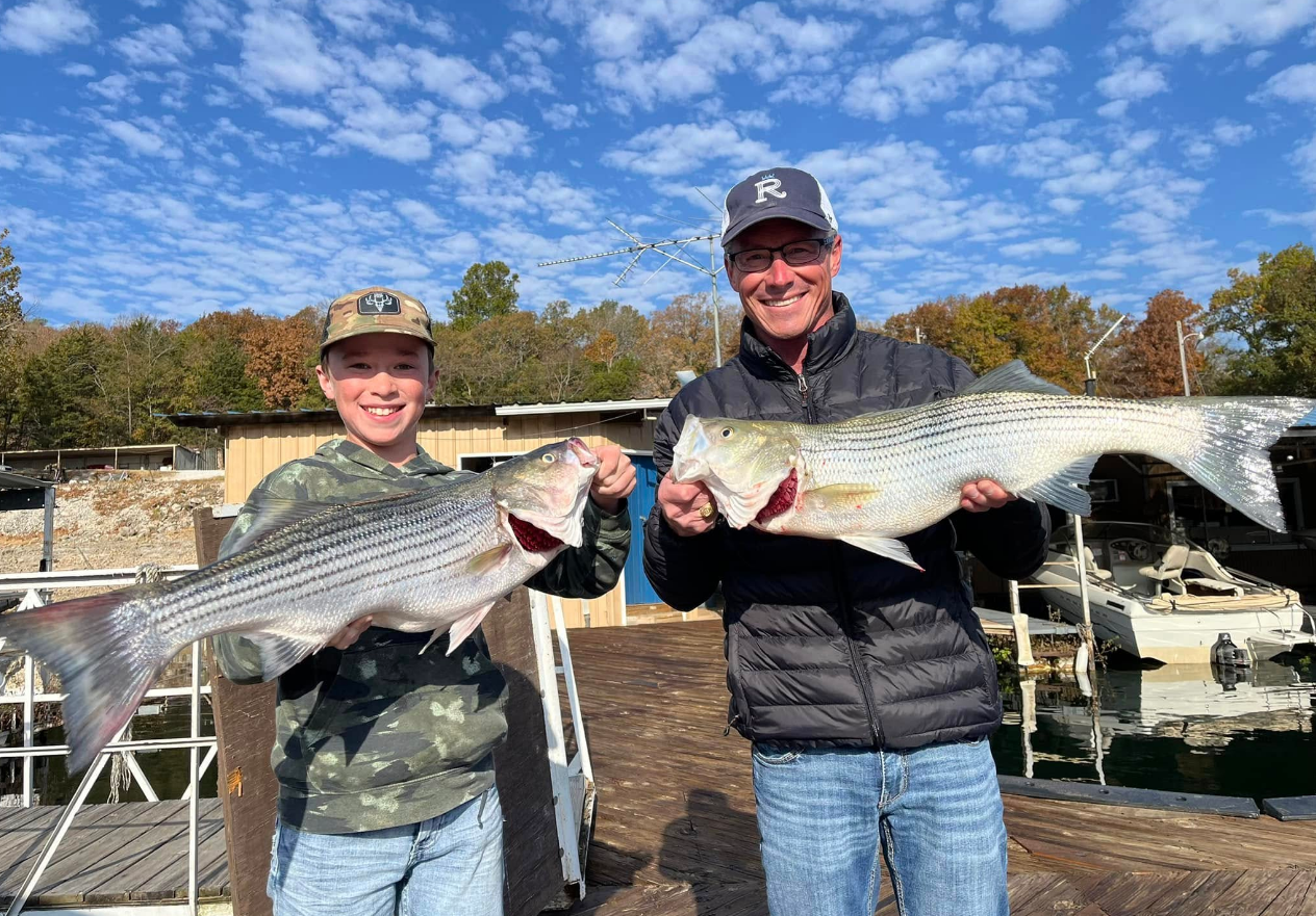 Two people holding large striped fish on a dock, smiling.
