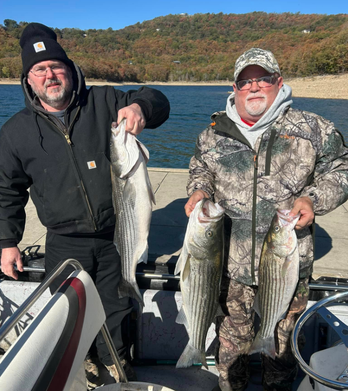 Two men on a boat show off striped bass they caught, near a lake with fall foliage.