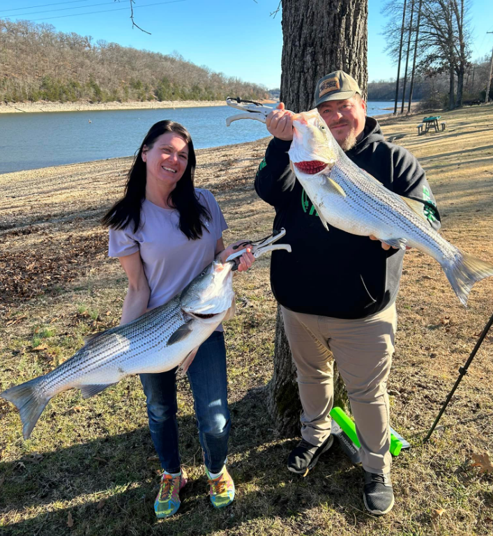 A woman and man smiling, holding up large striped fish they caught near a lake.