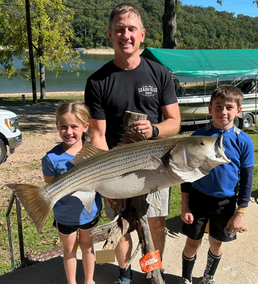 Man and two children holding a large striped bass fish by a lake.