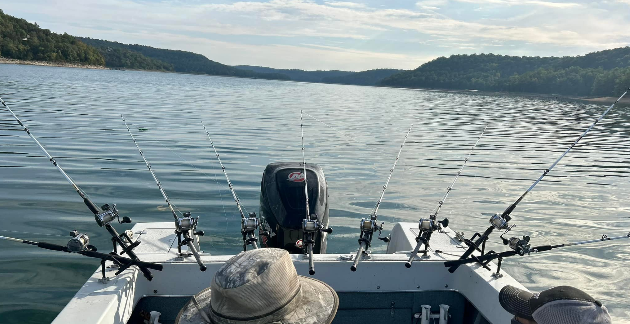 A person fishing on a boat with multiple fishing rods on a lake surrounded by forested hills.