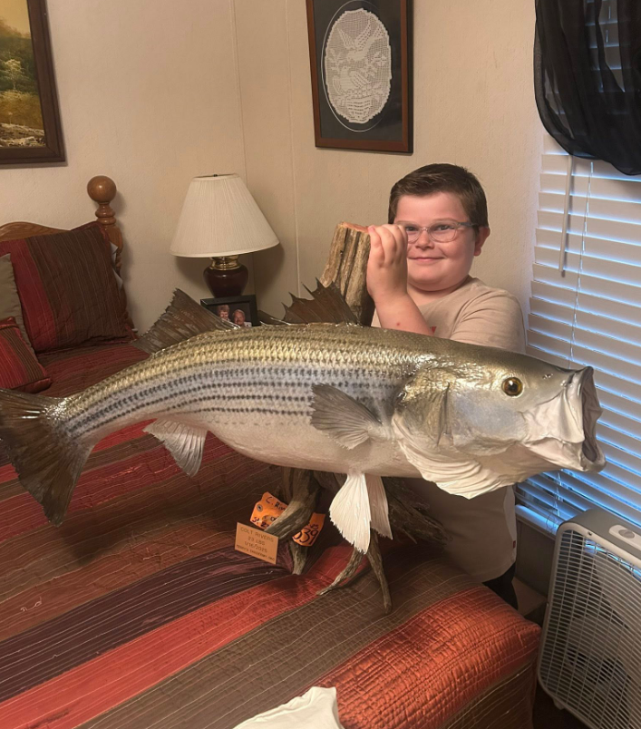 Boy holding a large mounted striped bass indoors, smiling.