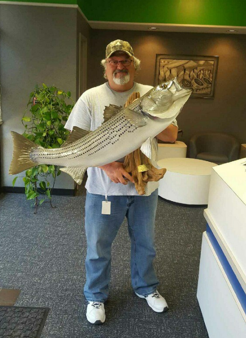 Man holding a large striped bass, posing on a boat with a scenic lake background.