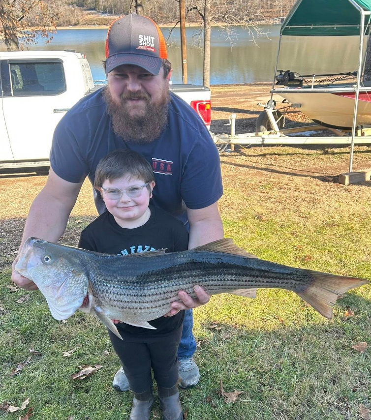 Man and child holding a large striped fish near a lake; they are smiling.
