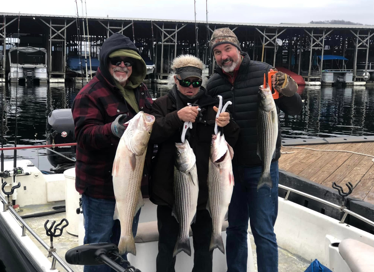 Three people on a boat display striped bass they caught, smiling by a marina.