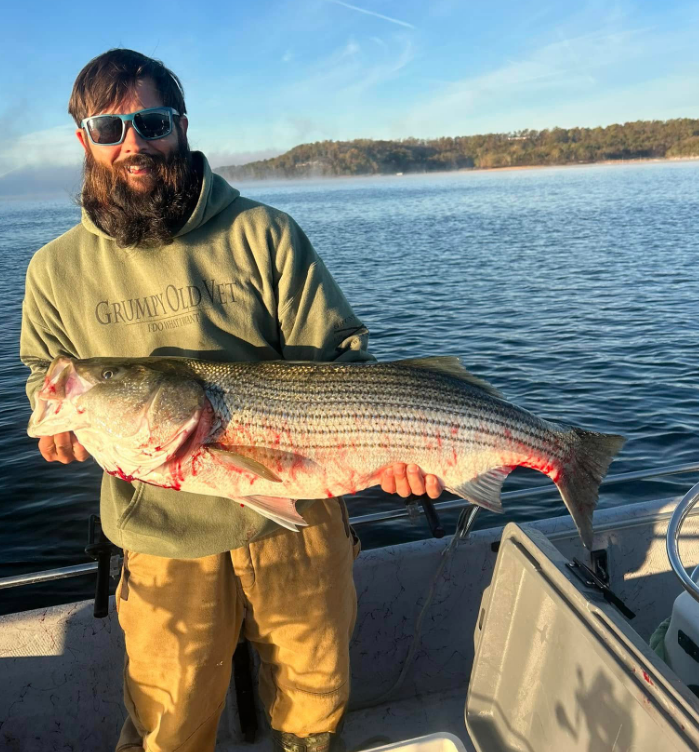 Man holding large striped bass on a boat, wearing sunglasses and a green hoodie, foggy lake backdrop.