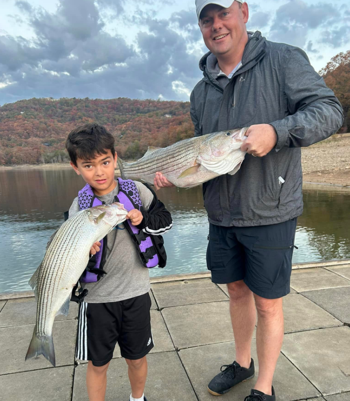 Man and boy holding striped bass on a dock by a lake. Boy wears a life vest, both smile.