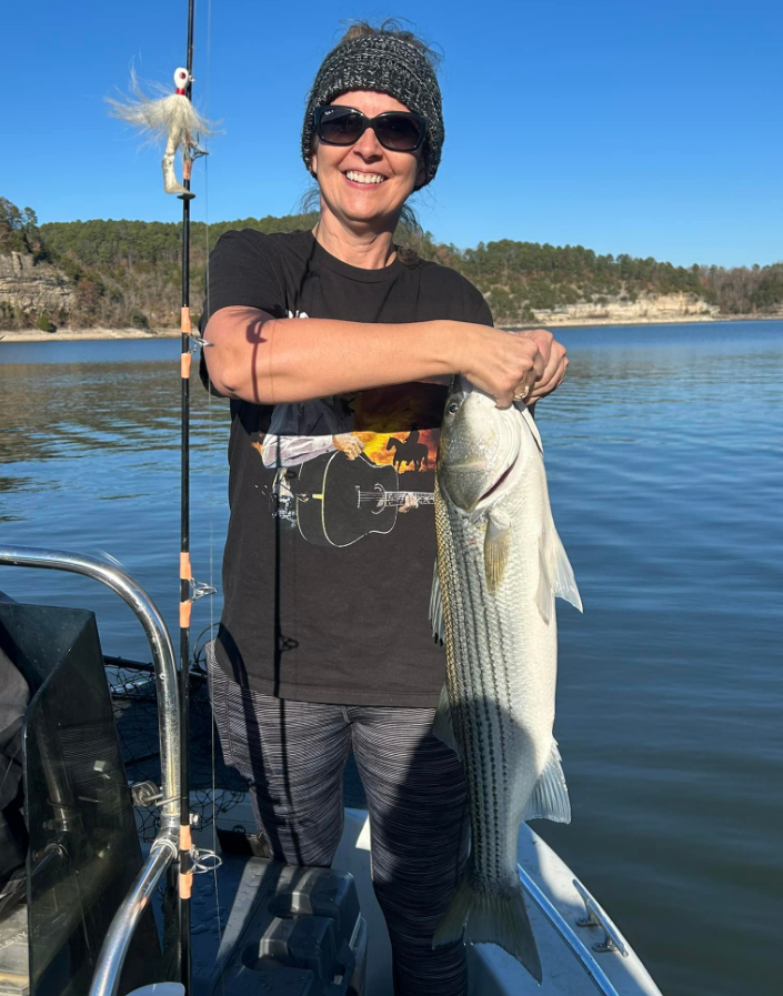 Woman smiles, holding a striped fish on a boat. Fishing rod visible. Water, sky, and trees in background.
