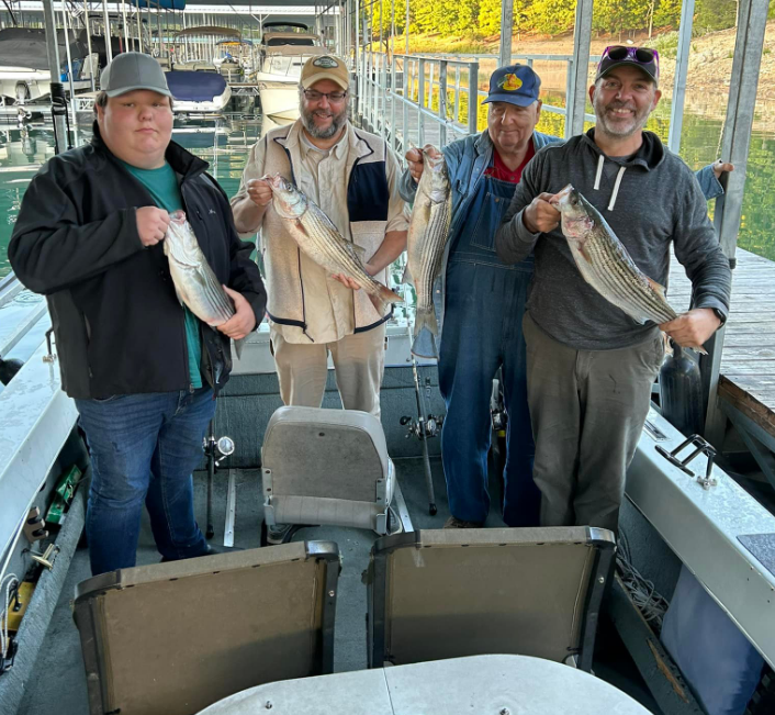 Four men on a boat, holding striped fish, smiling. Dock in background.