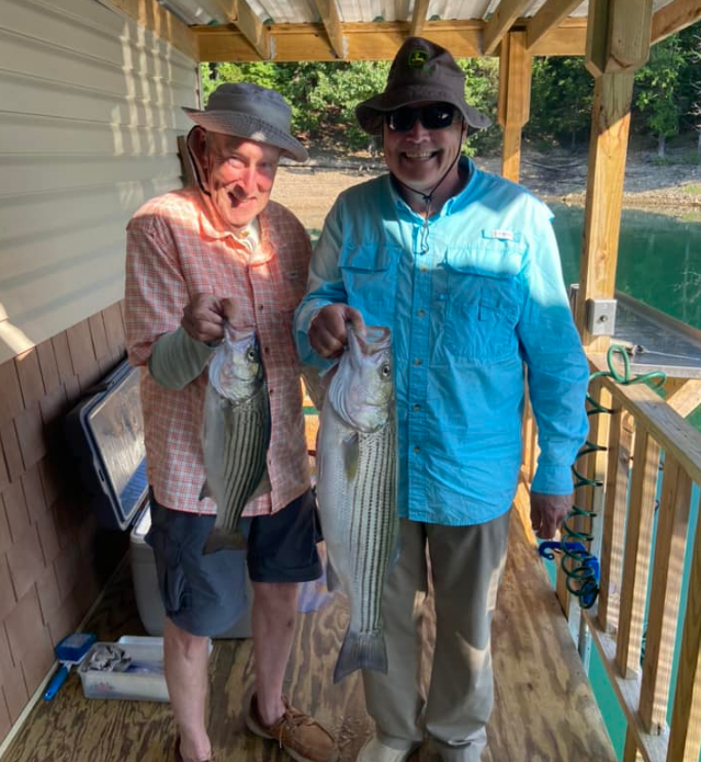 Two men holding striped fish on a wooden porch. One wears blue, the other orange, smiling.