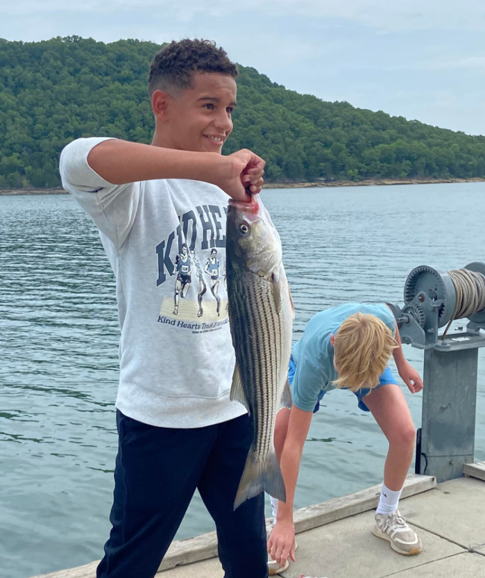 Boy holding a striped fish on a dock, another boy looking at it; lake and hills in the background.