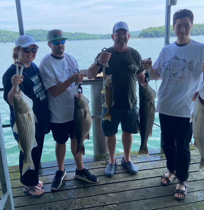Four people on a dock holding up fish they caught; lake in the background.
