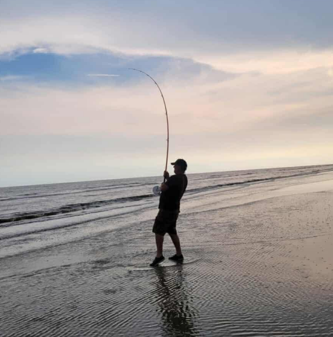 Man fishing on a beach with a bent rod under a cloudy sky.