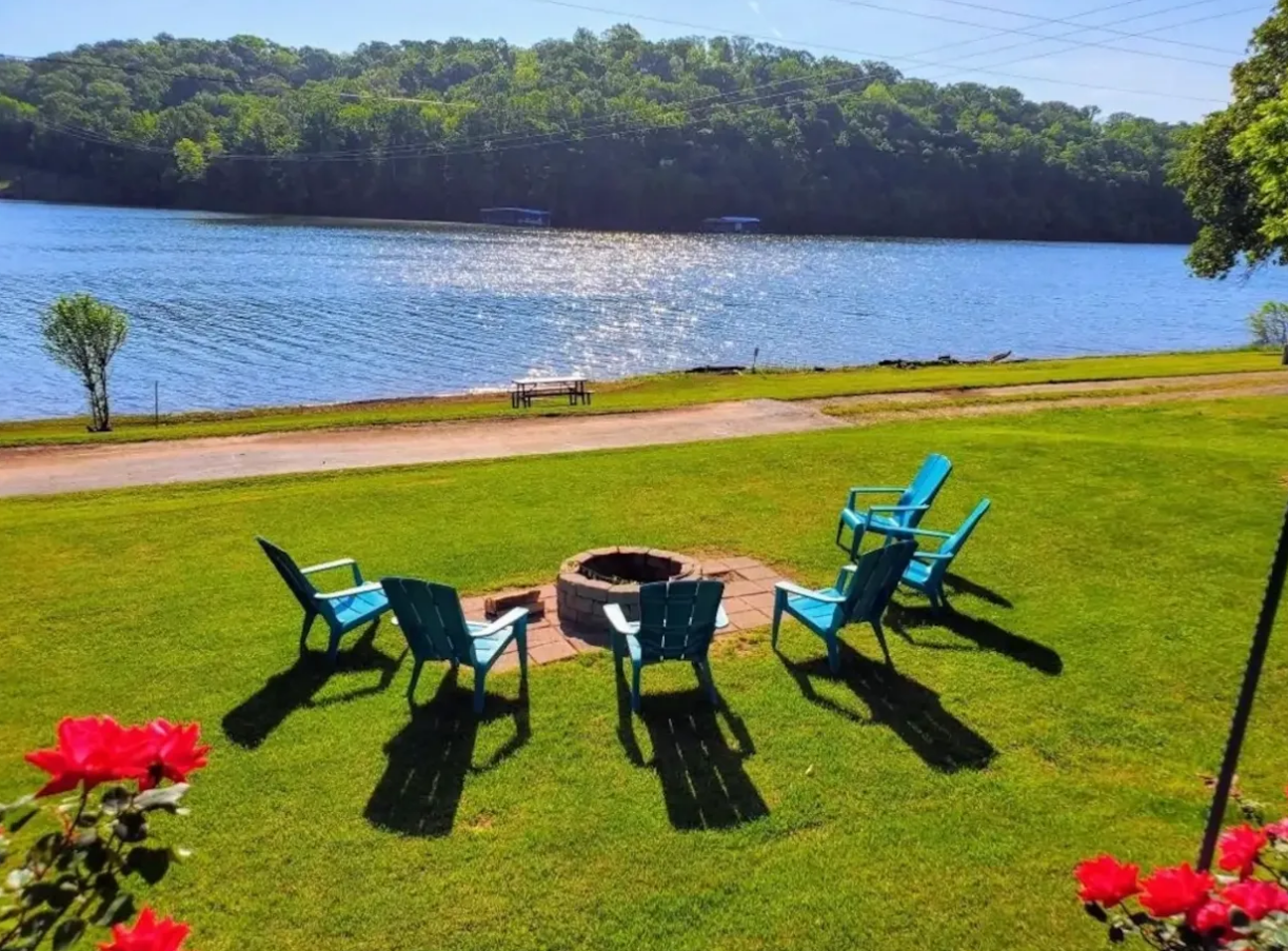 Fire pit with blue chairs on a grassy lawn overlooking a lake with trees.