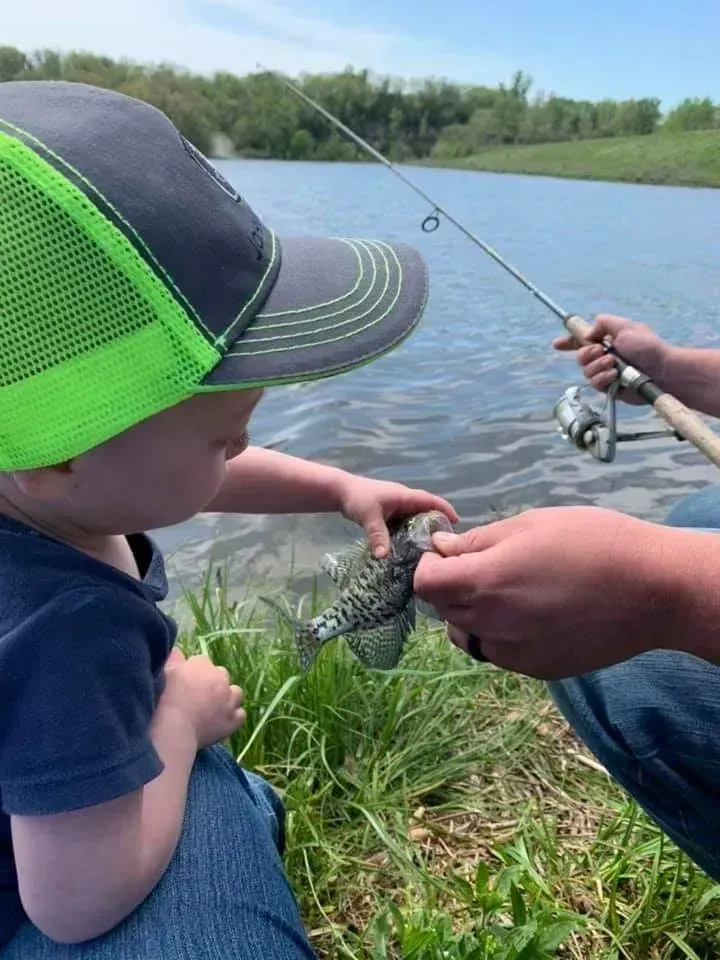 Child examining a fish held by an adult near a lake. Child wears a green and blue cap.