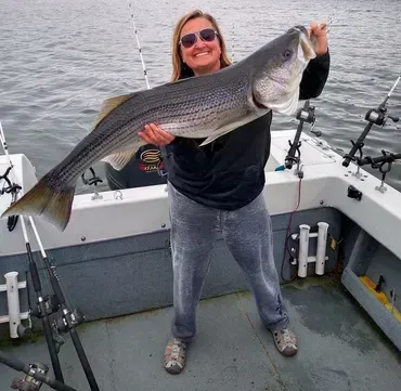 Woman on boat holding large striped bass. Fishing rods visible, overcast sky.