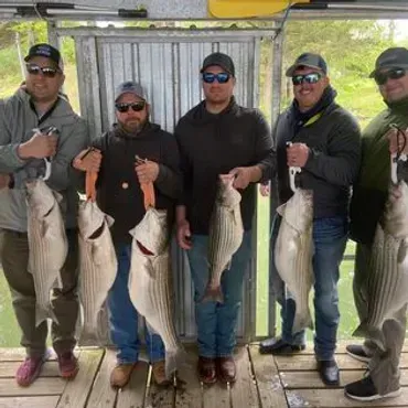 Five people holding up striped bass they caught, standing on a dock, cloudy sky.