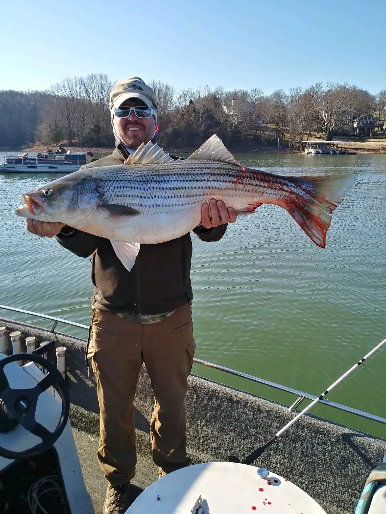 Man holding a large striped bass on a boat, water in background. Sunny day.