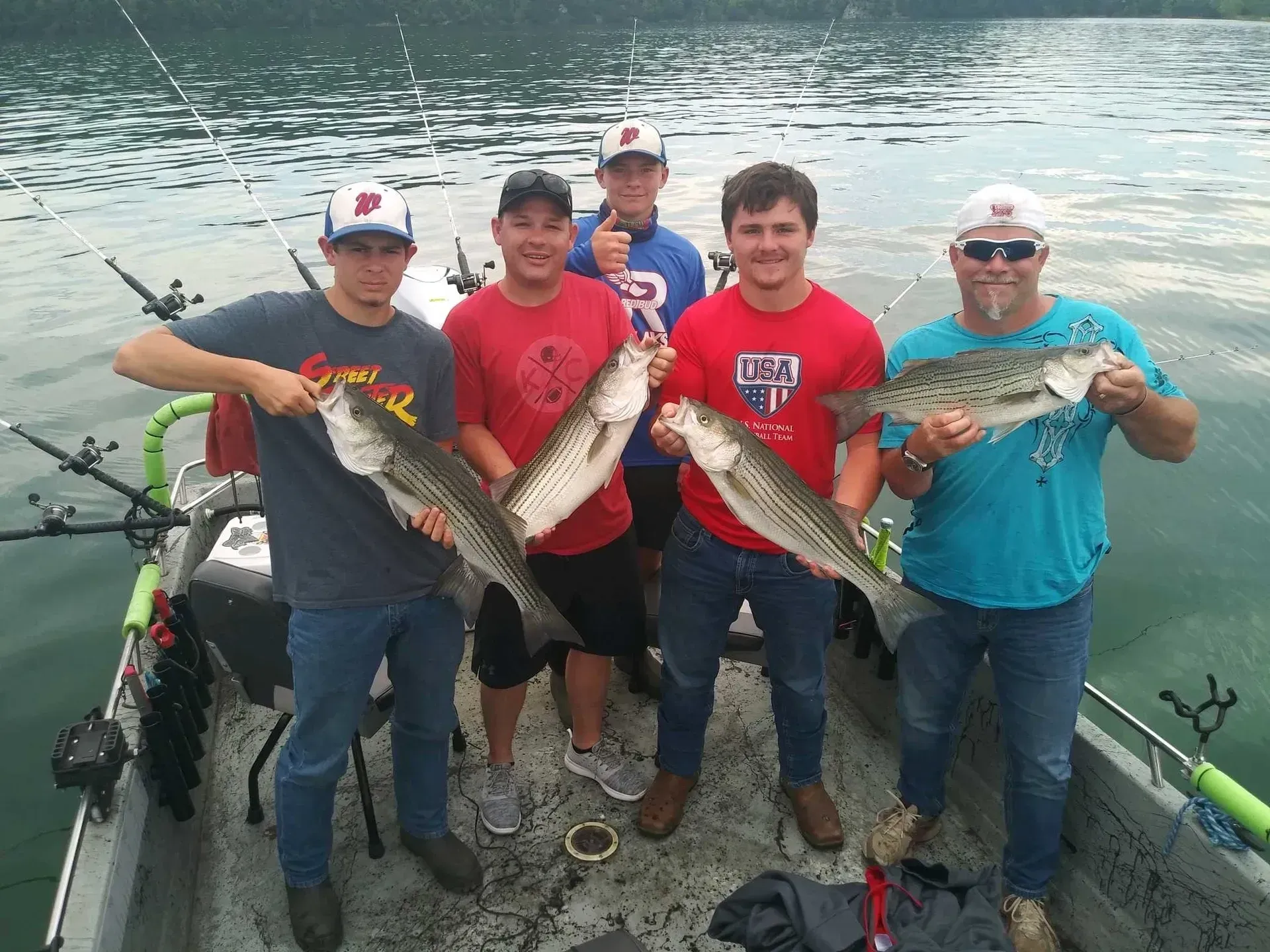 Five people on a boat display striped fish they caught. Cloudy water, overcast sky.