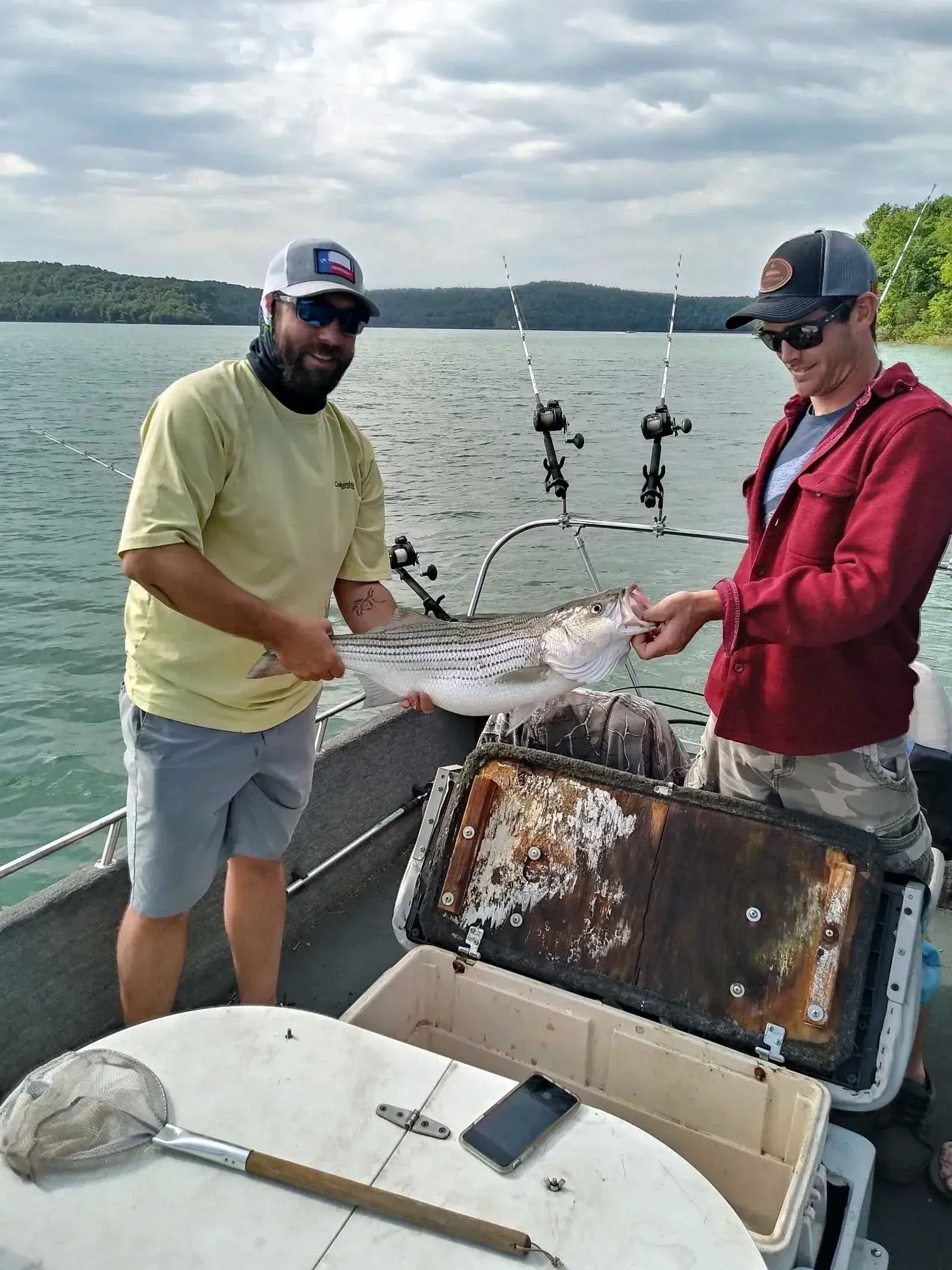 Two men on a boat holding a fish, lake in background. One man is using a net.