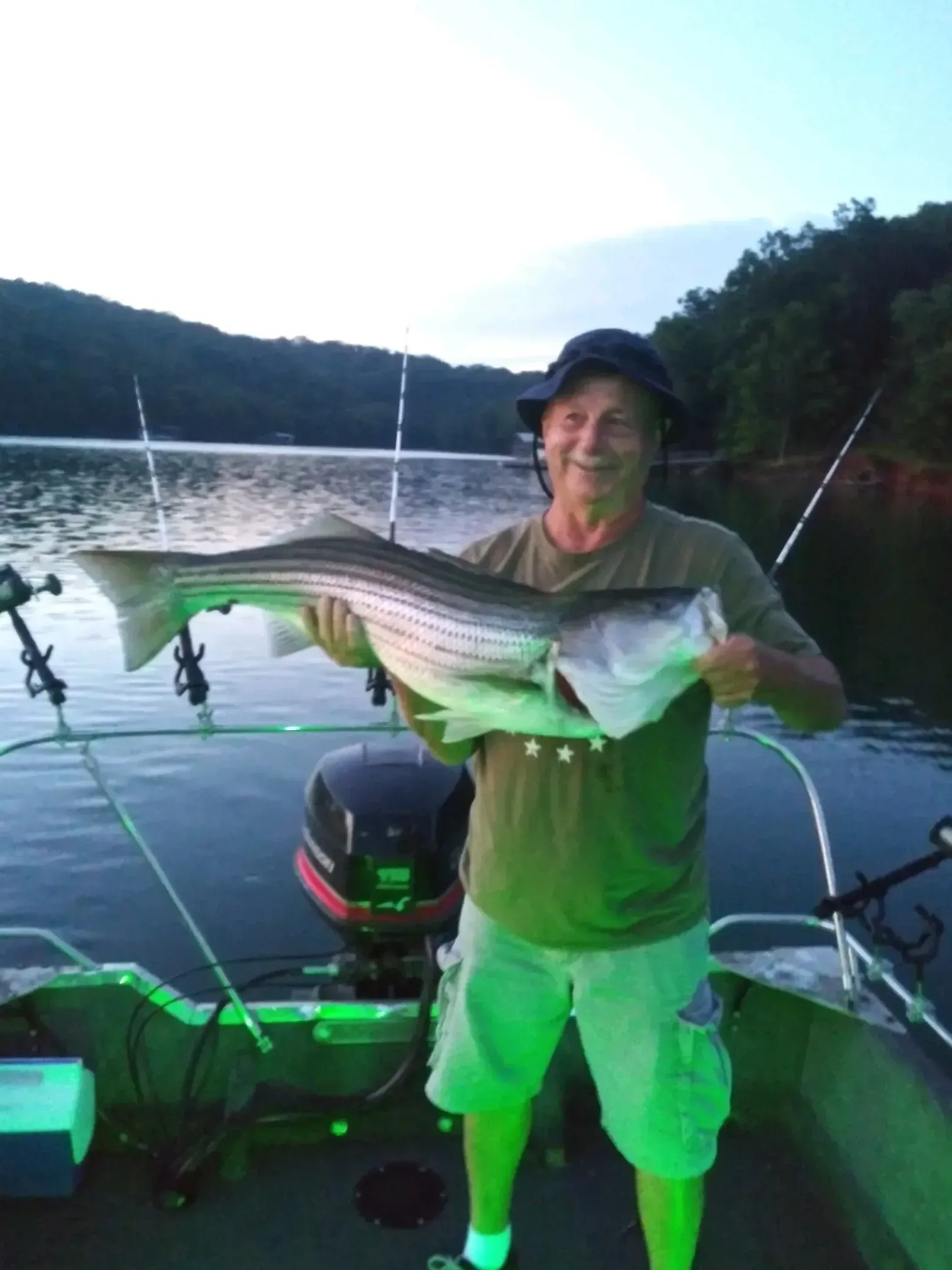 Man holding a large striped fish on a boat; lake and trees in the background, night.