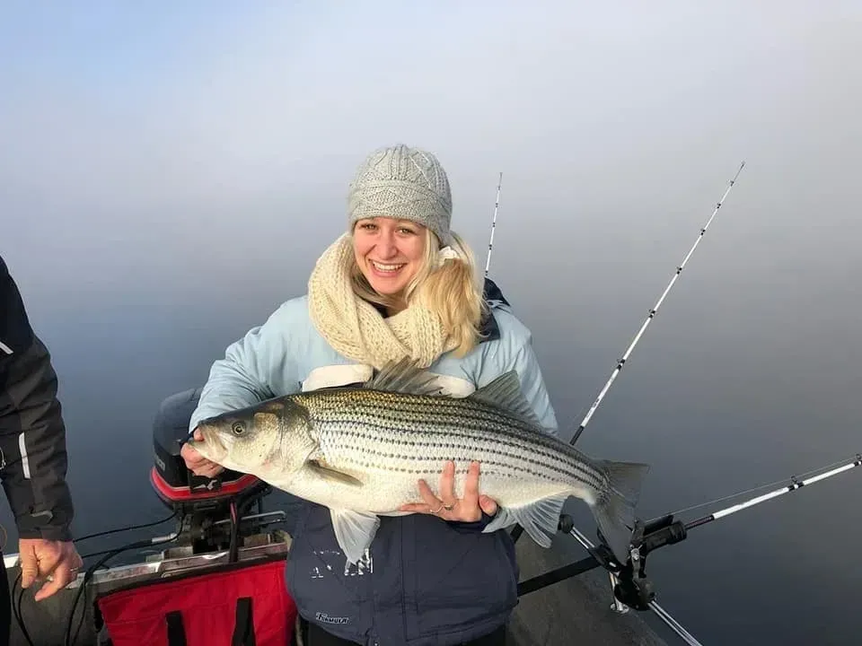 Woman on a boat holding a large striped fish; smiling near foggy water.
