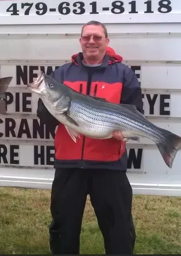 Man holding a large striped fish outdoors, wearing a red and blue jacket, with a business sign in the background.