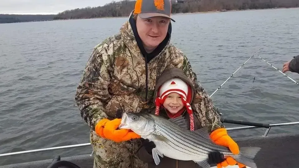 Man and child on a boat holding a striped fish. Child is smiling. Both are bundled up in winter gear. Lake in background.