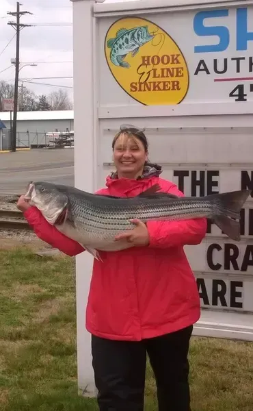Woman holding large striped bass in front of a sign for a fishing shop.