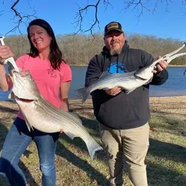 A woman and a man holding up two striped fish they caught near a body of water.