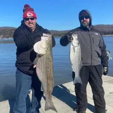 Two men holding up striped bass they caught, on a dock, by a lake.