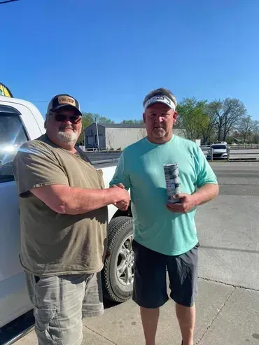 Two men shaking hands outside, holding a can of beverage near a white truck, sunny day.