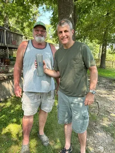 Two men outside holding a silver object. One wears a green shirt and shorts, the other a tank top, shorts, and a hat.