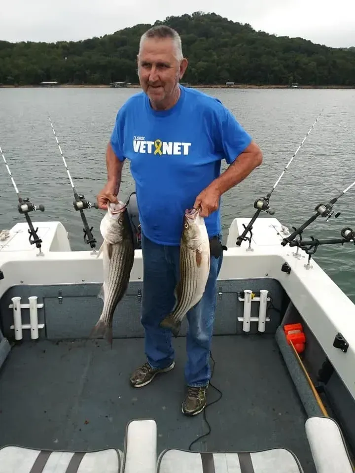Man on boat holds two fish; lake and trees in background. He wears a blue shirt and smiles.