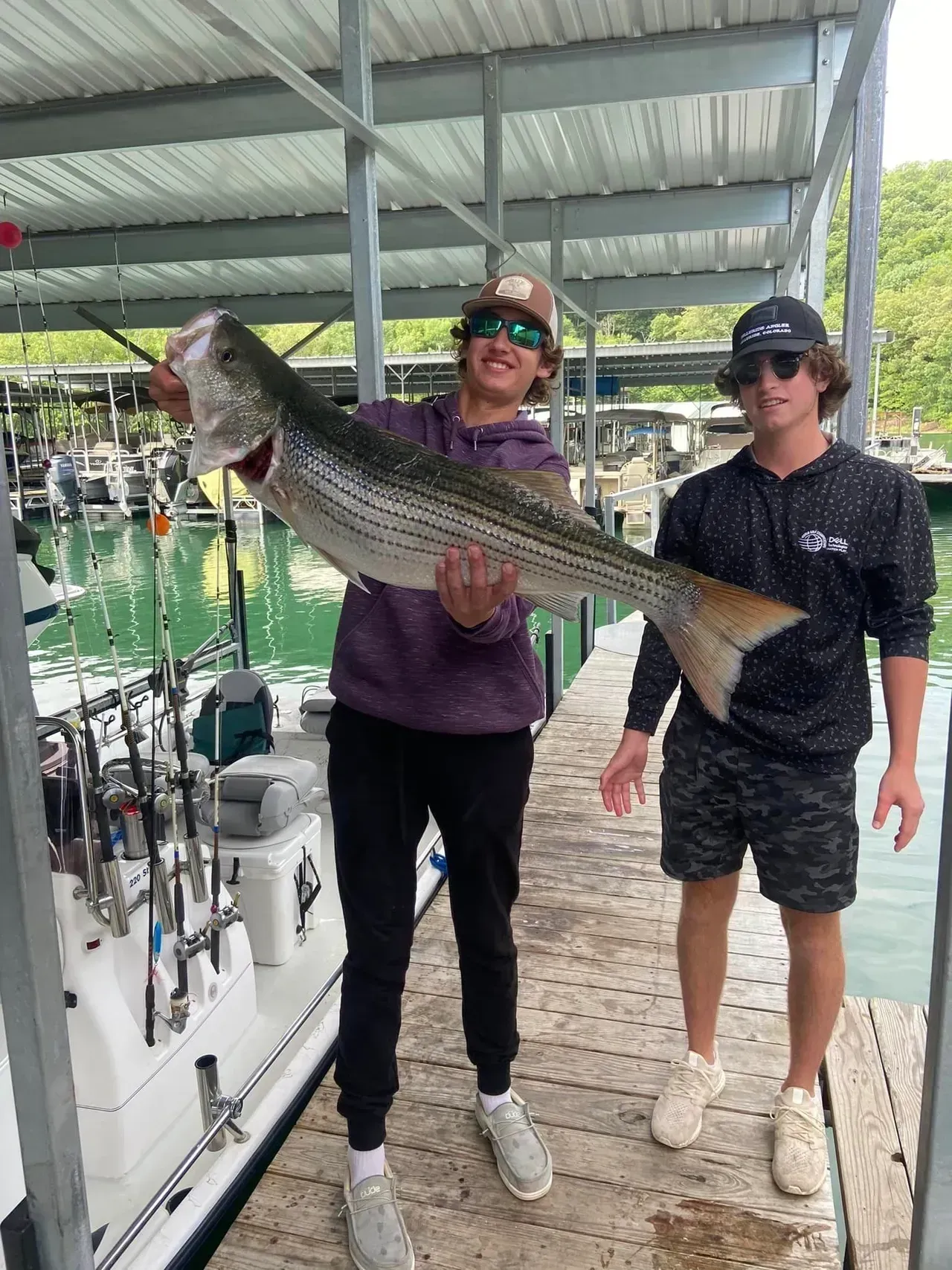 Two men holding a large striped bass on a dock. The fish is silver and black. Green water and boats are in the background.