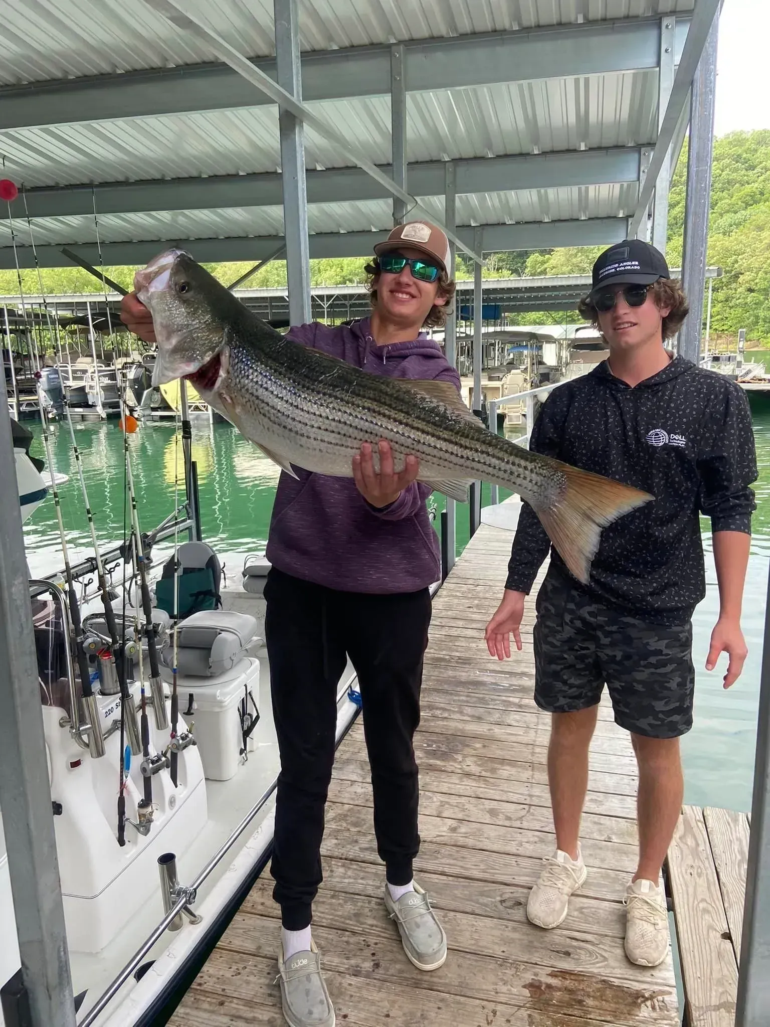 Two people holding a large striped fish at a dock with fishing rods and a boat.