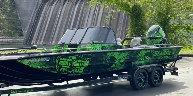 A green and black fishing boat on a trailer parked in front of a dam.