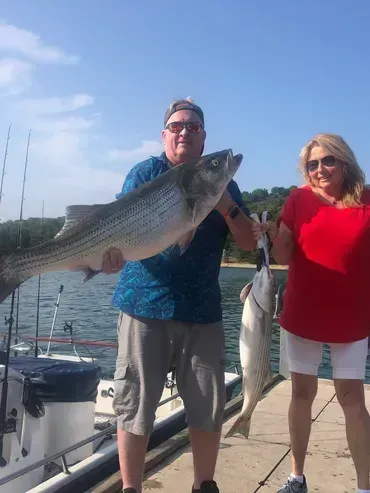 Man and woman on a dock holding up large fish, blue water in background. Bright sunny day.