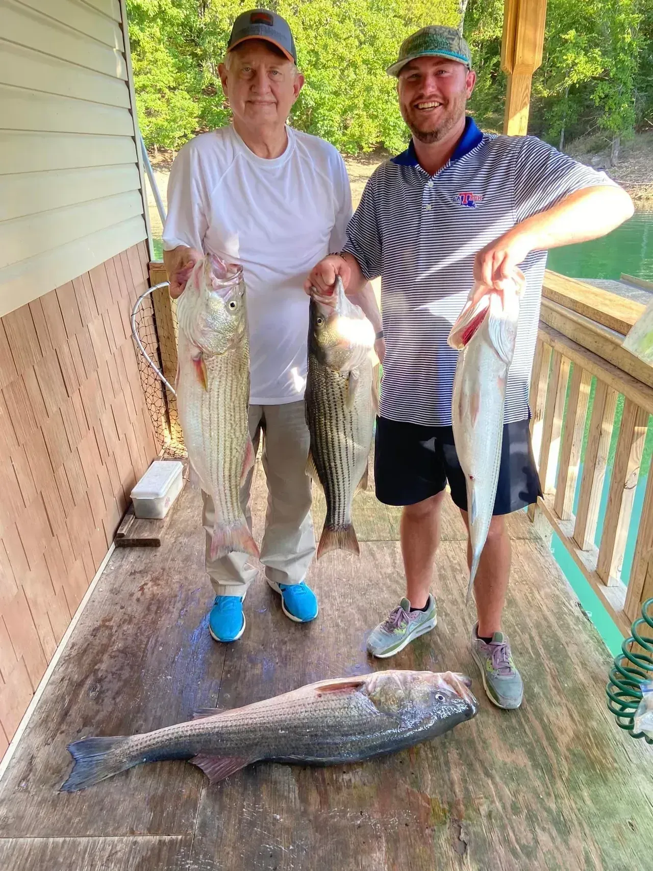 Two men holding striped bass on a porch, with one large fish on the floor; lake view background.